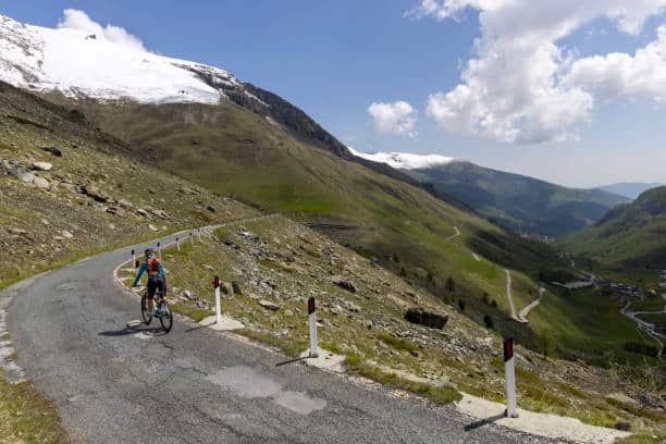 The road curves through a high mountain meadow towards snowcapped mouuntains