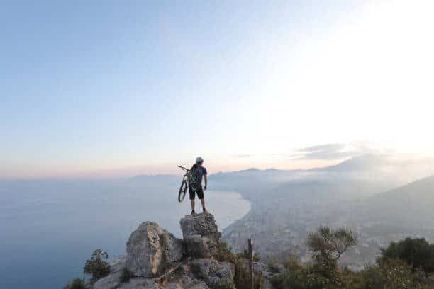 man on top of mountain holding bike viewing the city